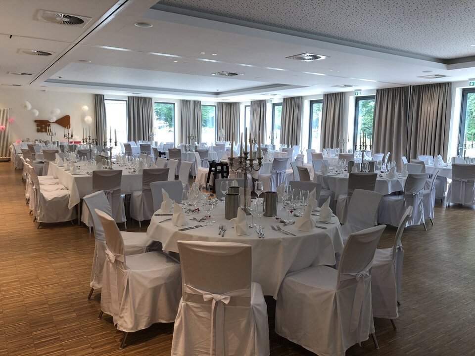 Ein geräumiger Bankettsaal mit weißen Tischen, weißen Tischtüchern und Silberbesteck ist für eine formelle Veranstaltung mit großen Fenstern und grauen Vorhängen eingerichtet.