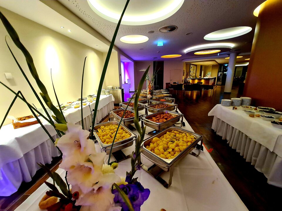 Ein langer Buffettisch mit weißen Tischdecken, silbernen Serviertabletts und einem floralen Tafelaufsatz ist in einem großen Raum mit hoher Decke, Kronleuchtern und einem Barbereich aufgebaut.