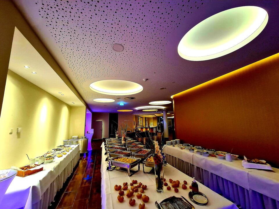 Ein Buffettisch mit weißen Tischdecken, silbernen Servierschalen und einer Vielzahl von Speisen steht vor einer gelben Wand, mit einer violetten Decke und einer roten Wand im Hintergrund.