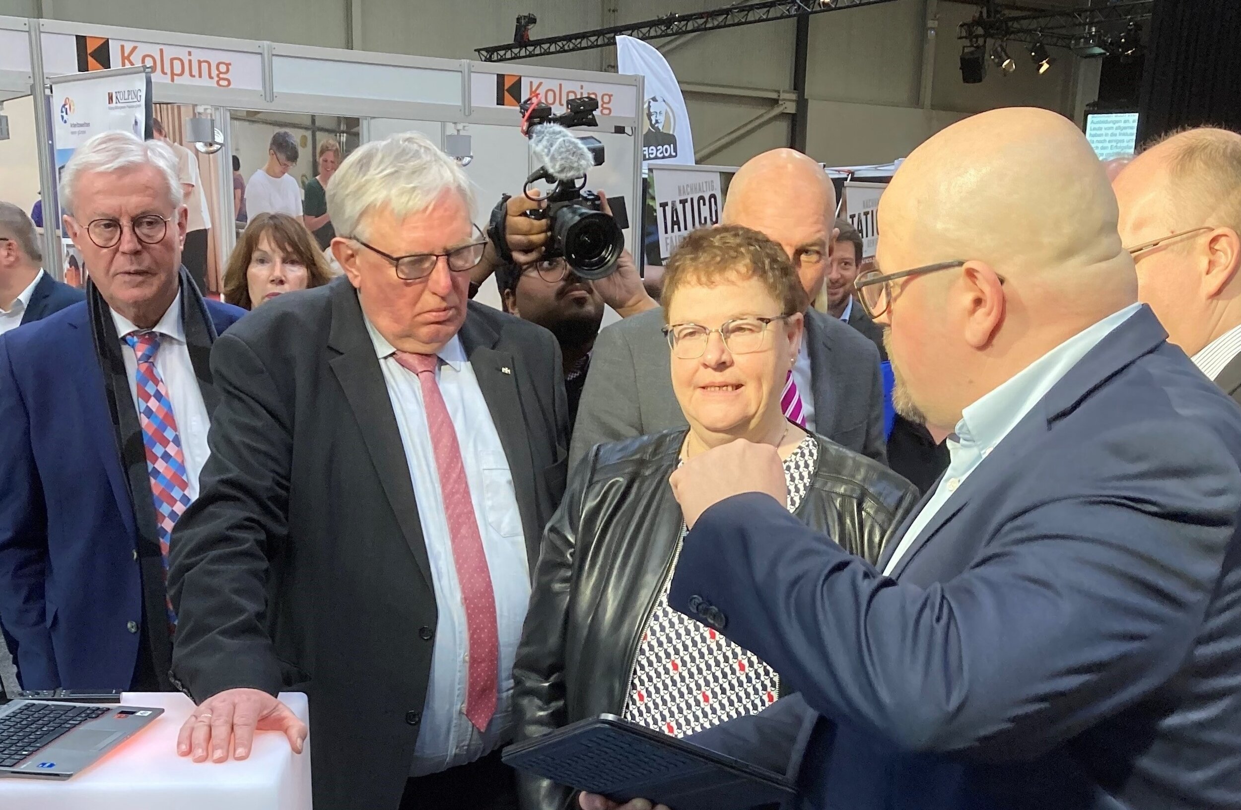 Auf einer Messe gestikuliert ein Mann in Anzug und Krawatte in Richtung einer Frau in einem schwarzen Jackett, während ein Mann in blauem Anzug und mit Brille beobachtet.