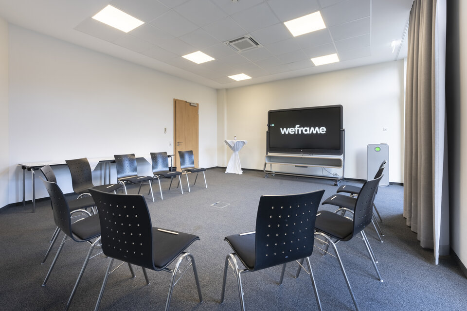 Ein moderner Konferenzraum ist mit einem großen Bildschirm, einem weißen Tisch, schwarzen Stühlen und einem weißen Podium ausgestattet, mit grauem Teppich und weißen Wänden.
