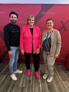 Drei in Schwarz, Rosa und Beige gekleidete Personen stehen vor einem roten Hintergrund mit dem Wort "Alexianer".