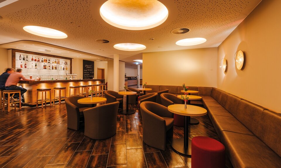 Der moderne Bar- und Loungebereich ist mit einer Holzbar, Barhockern, runden Tischen und einer langen Bank ausgestattet, die warm beleuchtet und mit einem roten Hocker versehen ist.