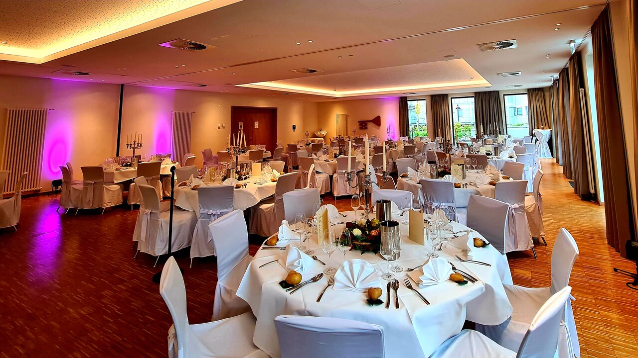 Ein großer Bankettsaal mit weißen Tischtüchern, Blumenschmuck und weißen Stühlen ist für eine formelle Veranstaltung eingerichtet, wobei ein großes Fenster und ein Kronleuchter ein warmes Licht verbreiten.