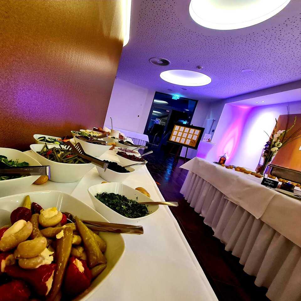 Auf einem Buffettisch in einem modernen Restaurant stehen verschiedene Gerichte, darunter Salate, Gemüse und Brot, auf einer weißen Tischdecke und vor einem lila Wandhintergrund.