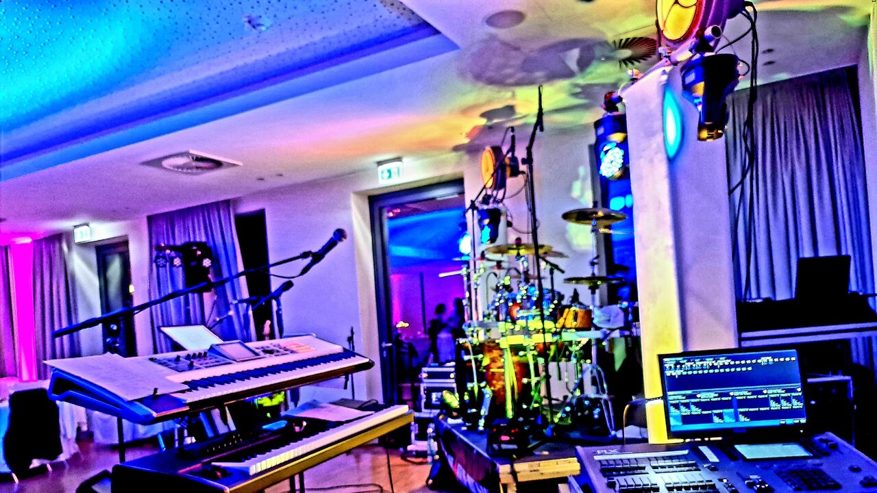 In einem schwach beleuchteten Raum befinden sich ein Keyboard, ein Schlagzeug und ein Computermonitor, im Hintergrund eine Bühne und Darsteller, die von blauen und violetten Lichtern angestrahlt werden.