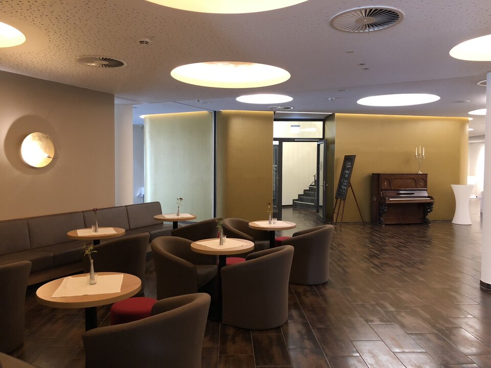 Das moderne Foyer mit Louge und Bar ist mit einem grauen Sofa, Couchtischen aus Holz und einem Flügel ausgestattet, über dem ein Kronleuchter und runde Leuchten hängen.