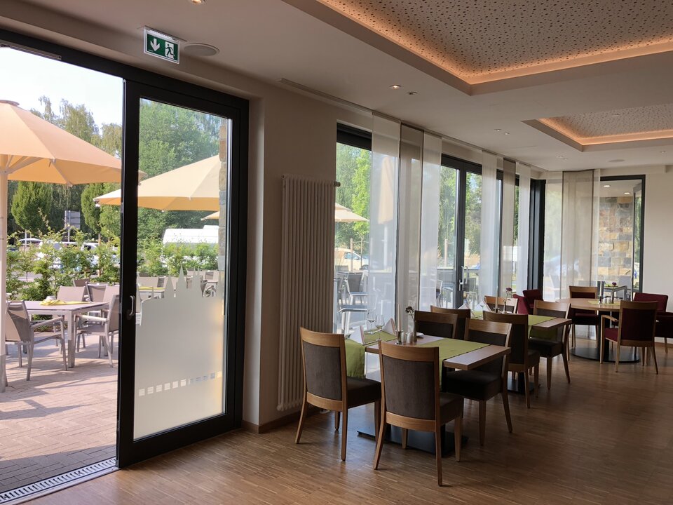 Das moderne Restaurant verfügt über ein großes Fenster mit Blick auf einen Innenhof, Holztische und -stühle, eine grüne Markise.