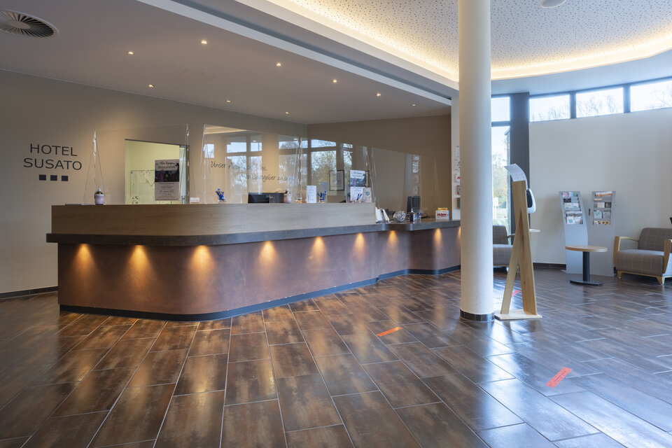 Eine moderne Hotelrezeption mit geschwungenem Holztresen und warmer Beleuchtung befindet sich in einer geräumigen Lobby mit hoher Decke, großen Fenstern und Blick auf Bäume.
