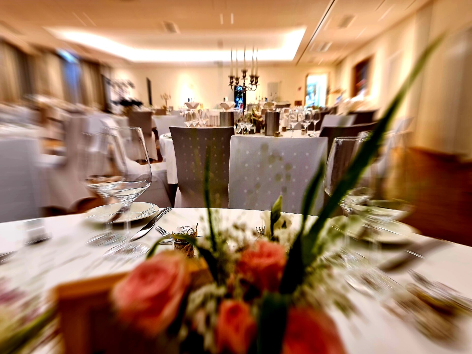 Ein Bankettsaal mit weißen Tischen, weißen Tischtüchern und Blumenschmuck ist für eine formelle Veranstaltung gedeckt, ein Kronleuchter und Kerzen verbreiten ein warmes Licht.