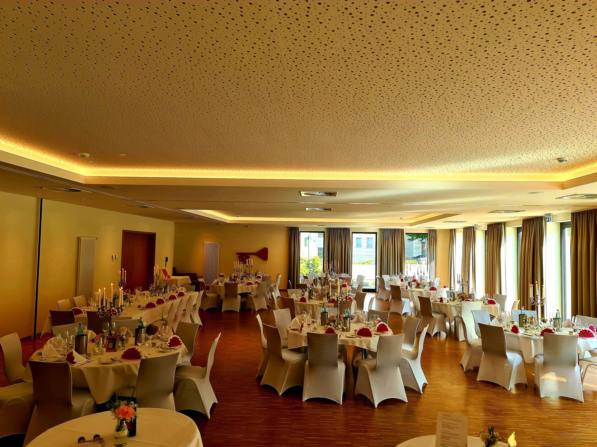 In einem geräumigen Bankettsaal mit hoher Decke, Holzboden und beigen Wänden stehen runde Tische mit weißen Tischtüchern, roten Servietten und Blumenschmuck, mit einem großen Fenster und einem Kronleuchter.