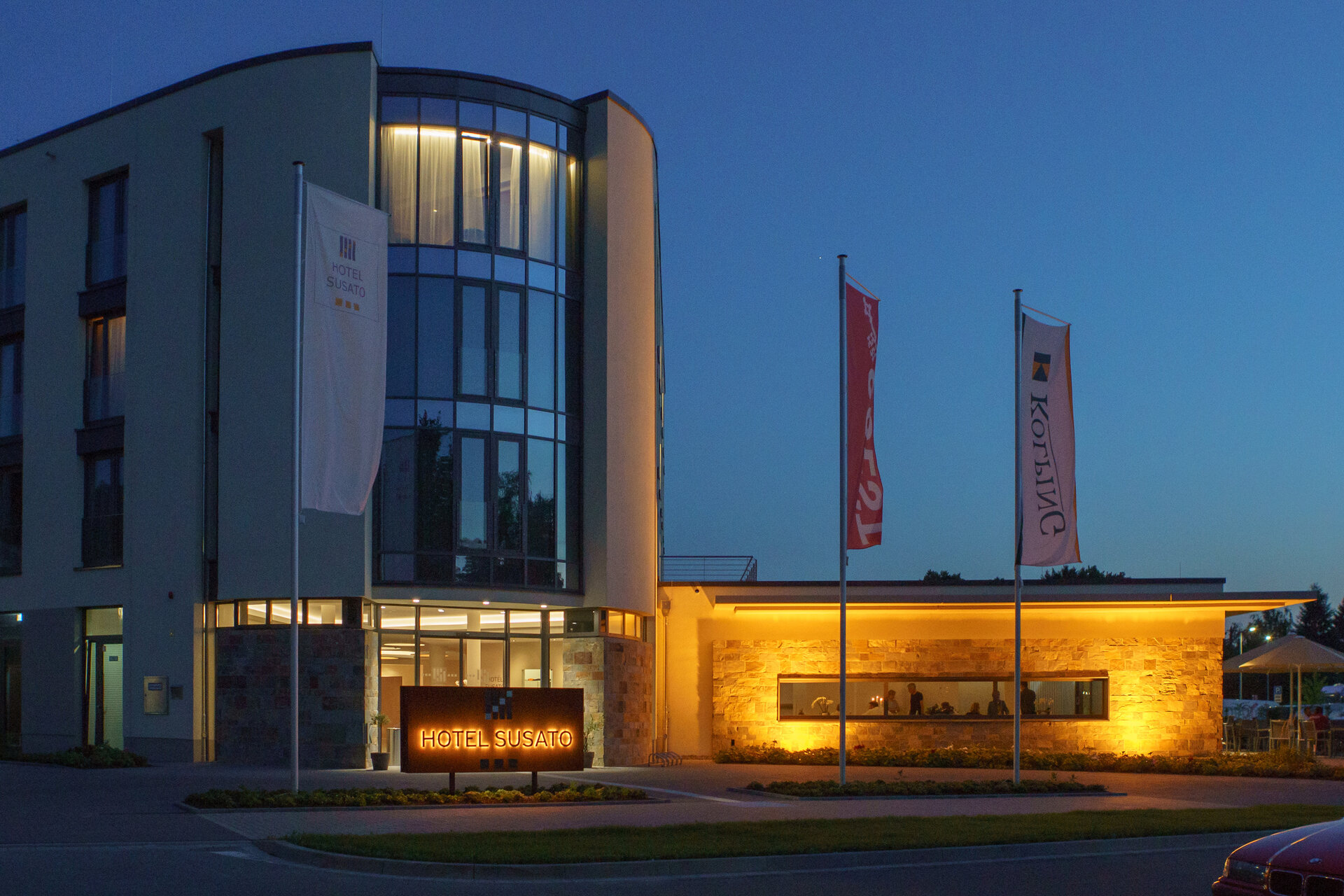 Das Hotel Susato, ein modernes Gebäude mit einer geschwungenen Fassade und großen Fenstern, ist nachts beleuchtet, und im Vordergrund flattern zwei Fahnen.