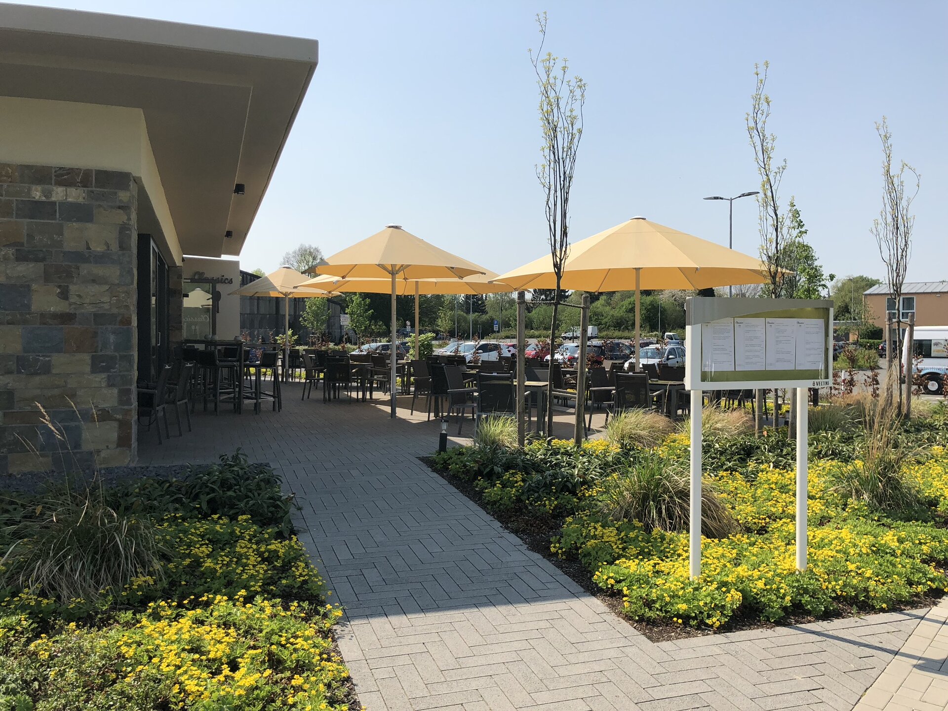 Ein modernes Restaurant mit einer Steinfassade und gelben Sonnenschirmen liegt in einem üppigen Garten, begleitet von einem weißen Schild und einem Parkplatz.