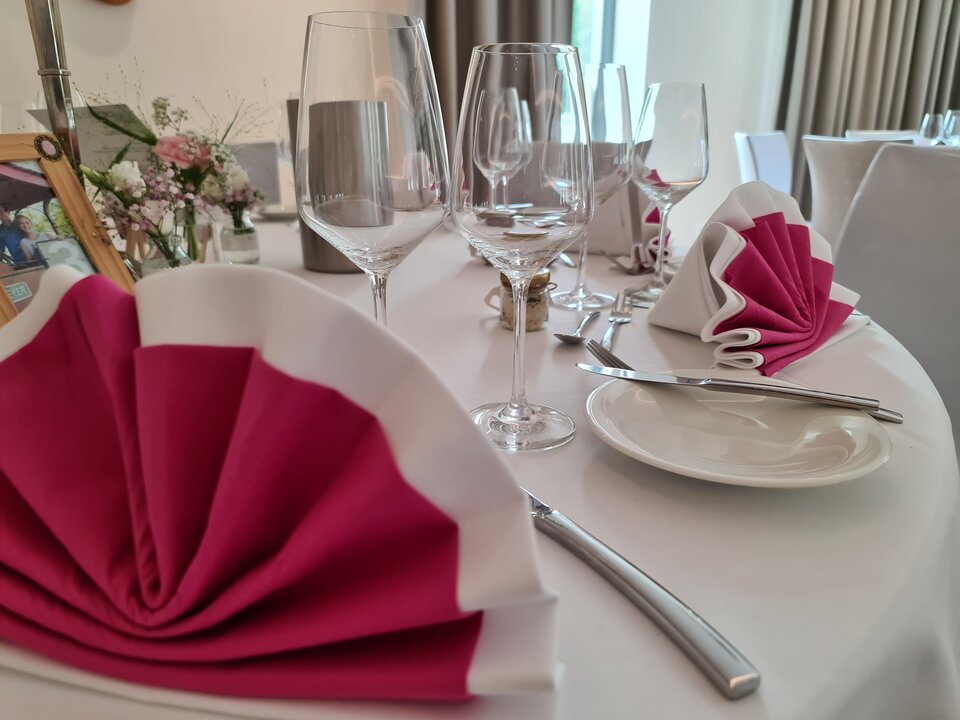 Ein weißes Tischtuch mit rosafarbener und weißer Serviette, Silberbesteck und Weingläsern ist für ein formelles Abendessen gedeckt, mit einer Blumenvase und Blick auf ein Fenster.