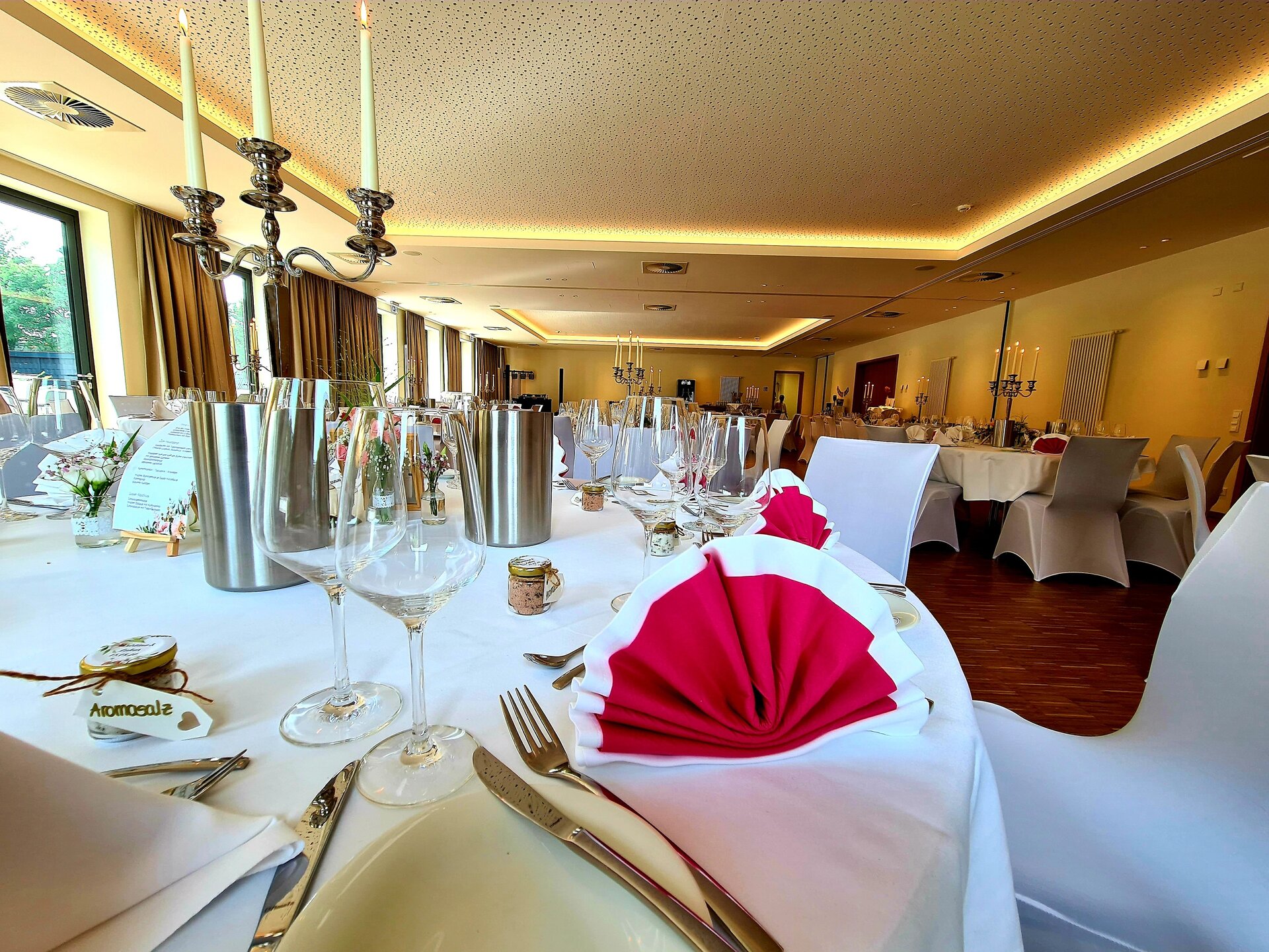 Ein runder Tisch in einem Bankettsaal ist mit weißem Tischtuch, roten Servietten, Silberbesteck und Weingläsern gedeckt und wird von einem Kerzenleuchter und einem Blumenschmuck begleitet.