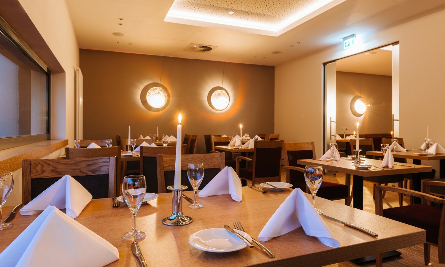 Das warm beleuchtete Restaurant ist mit Holztischen, weißen Tischtüchern und Silberbesteck ausgestattet. Ein großes Fenster und zwei runde Wandleuchten spenden ein sanftes Licht.